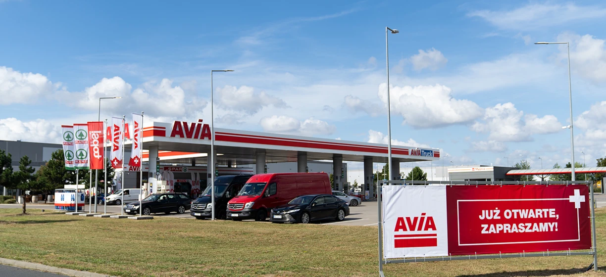 Lusterko boczne ciężarówki z reklamą AVIA, SPAR i SD Eibar na stacji benzynowej. Znak "AVIA Truck" z napisem po polsku "JUŻ OTWARTE, ZAPRASZAMY!" w tle.